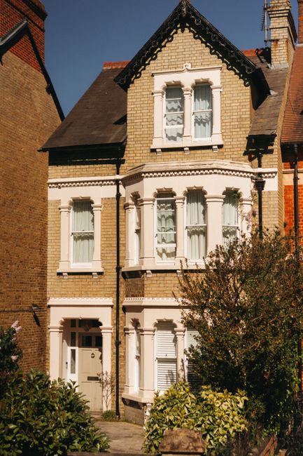An exterior view of a Victorian-style residential house with a brick facade, white decorative window frames, and three prominent front windows. The house has a sloped dark-colored roof with a chimney,