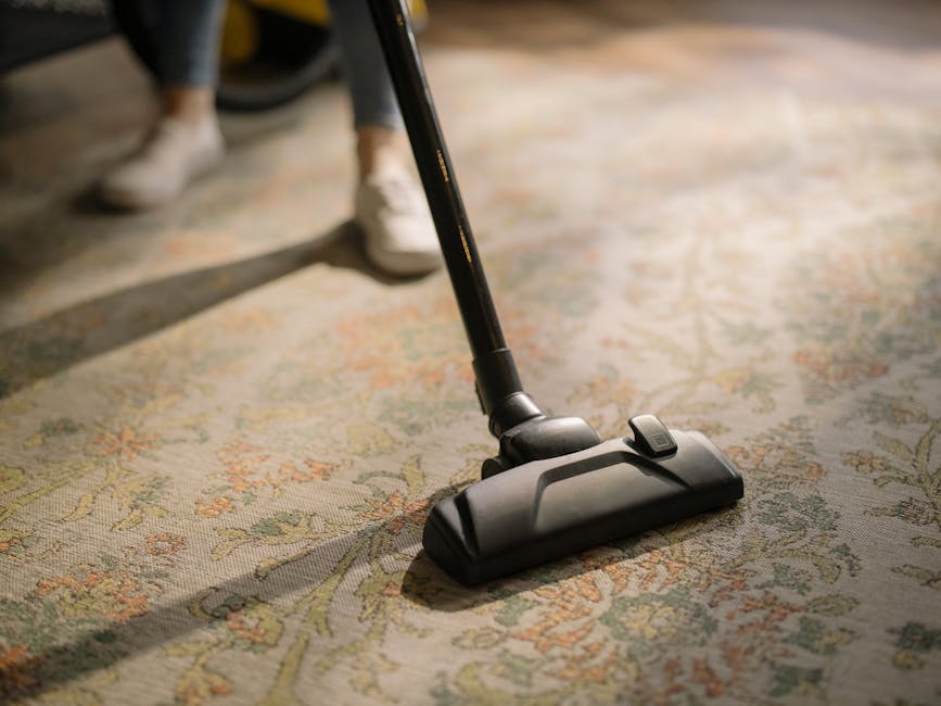 Close-up image of a vacuum cleaner head positioned on a patterned carpet within a room, with parts of a person's legs and grey shoes visible in the background. The carpet features a floral or ornate design with pastel colors, and the lighting suggests natural daylight. The scene emphasizes surface cleaning and maintenance, with the vacuum in the process of deep cleaning the residential textile flooring. This image illustrates professional carpet cleaning practices employed by Cleaners N7, a reputable UK cleaning company serving Tufnell Park and surrounding areas.